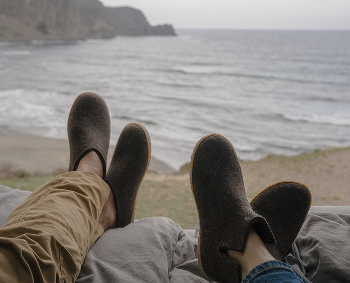 Two people relaxing with their feet up, looking out over a sandy beach and ocean waves under an overcast sky; the person on the left is wearing Chestnut, brown glerups slip-on, while the person on the right is wearing Chestnut, dark glerups boots, both with honey rubber sole. 
